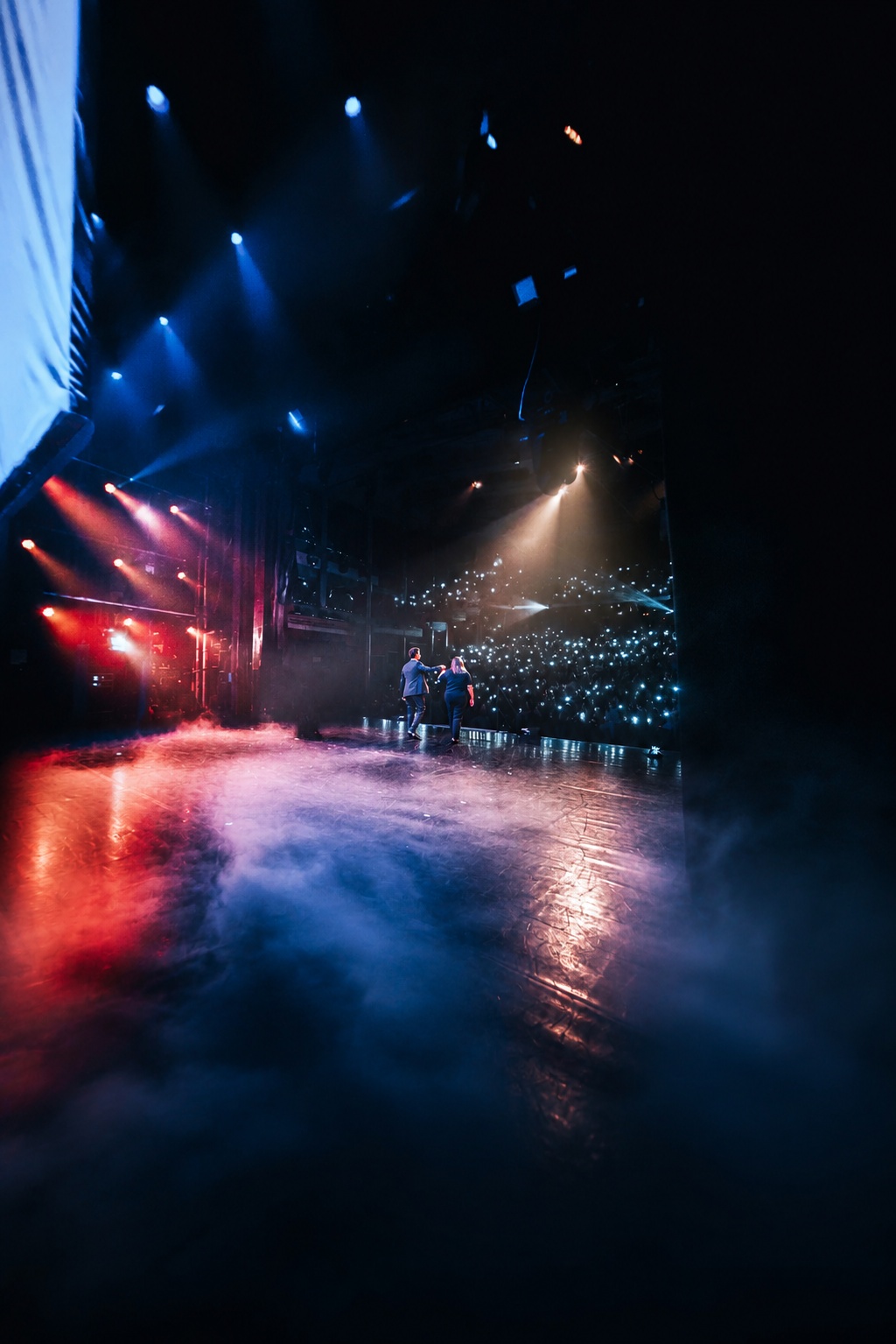 Jonas De Bruyn walking on stage with a spectator, blue and red lights, the audience holding their phone lights up